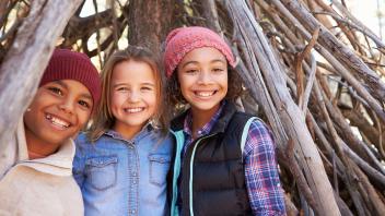 Three young kids standing in front of a fort made of sticks