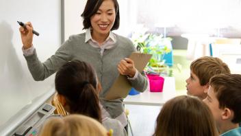 second grade teacher writing on whiteboard with students around her